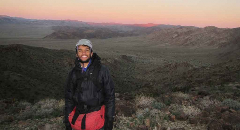 A person wearing a helmet smiles for the photo while standing high above a desert landscape below. They sky appears in shades of pink. 